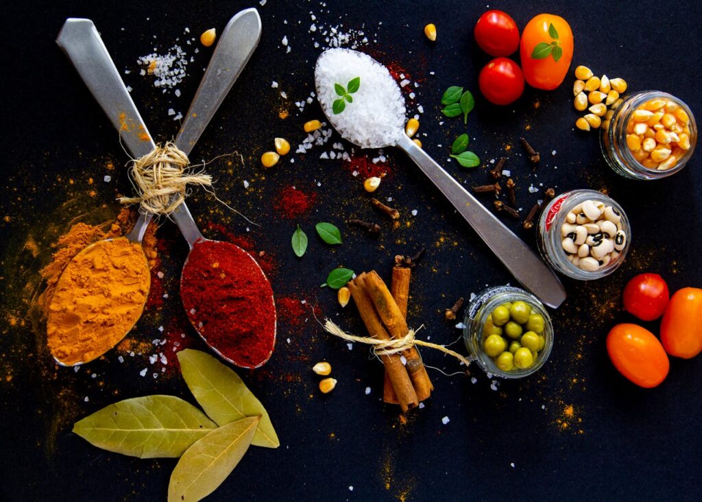 Top view of dry turmeric and paprika near bay leaves with cinnamon sticks and sea salt in spoons near jars with popcorn grains and olives with ripe colorful cherry tomatoes